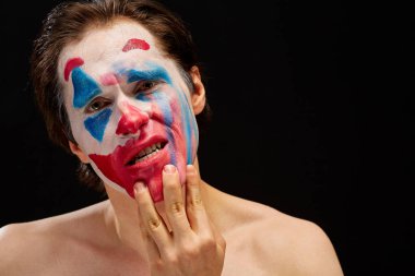Genç bir adam tek eliyle yüzüne palyaço makyajı sürer, çıplak omuzlarıyla, duyguları ifade eder, siyah bir arka planda.