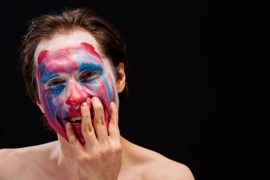 Genç bir adam tek eliyle yüzüne palyaço makyajı sürer, çıplak omuzlarıyla, duyguları ifade eder, siyah bir arka planda.