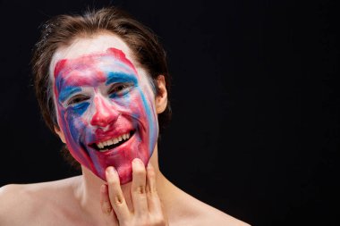 Genç bir adam tek eliyle yüzüne palyaço makyajı sürer, çıplak omuzlarıyla, duyguları ifade eder, siyah bir arka planda.