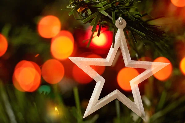 Ağaçta asılı duran beyaz yıldızlı Noel oyuncağı yeşil iğneler ve renkli bokeh