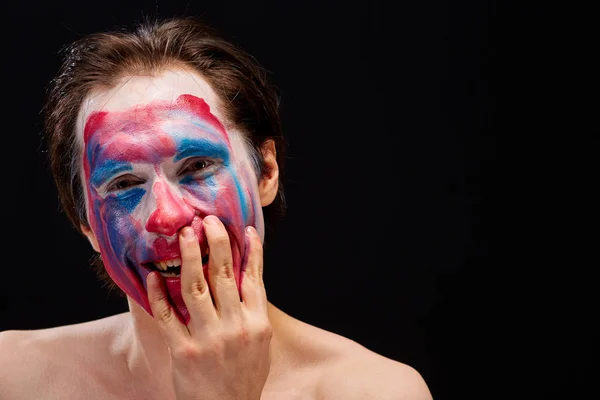 Genç bir adam tek eliyle yüzüne palyaço makyajı sürer, çıplak omuzlarıyla, duyguları ifade eder, siyah bir arka planda.