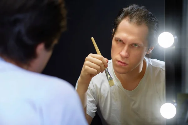 Oyuncu, karanlık bir odada beyaz bir tişörtle aynanın karşısına oturur ve yüzüne makyaj yapmak için elinde bir fırça tutarak yansımasına bakar.