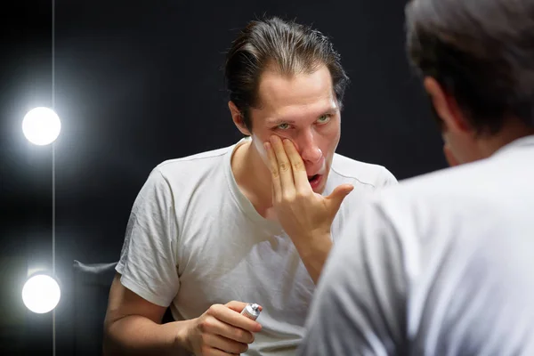 Aktör, bir elinde krem tüpü olan karanlık bir odada beyaz bir tişörtle aynanın karşısına oturur ve diğer elinde kremi yüzüne sürer.