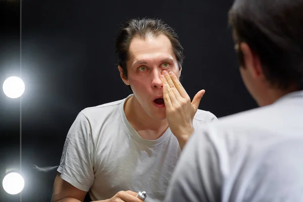 Aktör, bir elinde krem tüpü olan karanlık bir odada beyaz bir tişörtle aynanın karşısına oturur ve diğer elinde kremi yüzüne sürer.