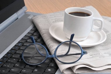 Bir fincan kahve gazetenin üstünde, bardakların ve dizüstü bilgisayarın yanında.