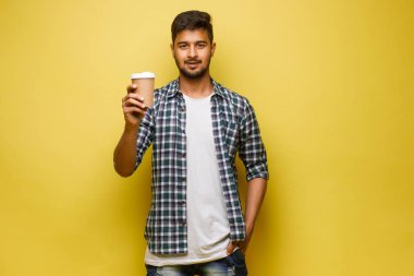 Genç Hintli esmer öğrenci elinde kahve fincanıyla sarı arka planda duruyor.