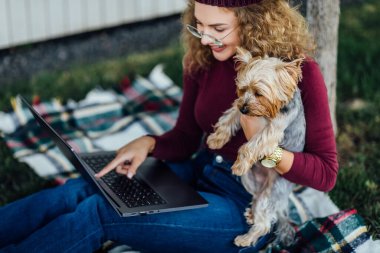 Öğrenci kadın battaniyenin üzerinde oturuyor ve Yorkshire teriyeri köpeğiyle piknik yapıyor. Bilgisayara bak..
