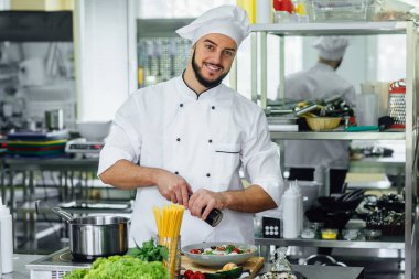 Profesyonel sakallı erkek, şef profesyonel bir mutfakta eğleniyor, salata ve spagetti hazırlıyor..