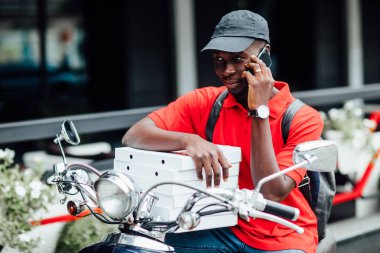 Genç Afrikalı adamın portresi motosikletle siparişi kabul ediyor. Elinde pizza kutusuyla motosikletine biniyor..