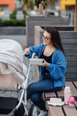 Anne bebek arabasında bebek arabasıyla yürüyor, kadın plastik kutu tutuyor ve bebeğini besliyor. Öğle yemeği vakti..