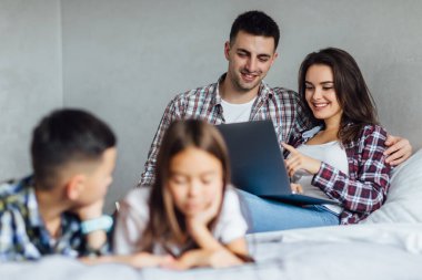 Yatak odasında yatıp dizüstü bilgisayar kullanan iki mutlu ebeveyn. Çocuklarla hafta sonu.