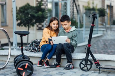 Küçük çocuklar motosiklet ve kaykayla sokakta yürürken dijital dizüstü bilgisayar kullanıyorlar..