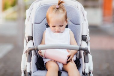 Pembe bir şişeyle bebek arabasında oturan küçük kız, bebek arabasıyla yürüyüşe çıkan bebek, yazın dışarıda gezen çocuk..