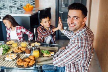 Mutlu baba ve oğul portresi günlük giysiler içinde el ele veriliyor, müsli yerken gülüyorlar evdeki ahşap masada birlikte..
