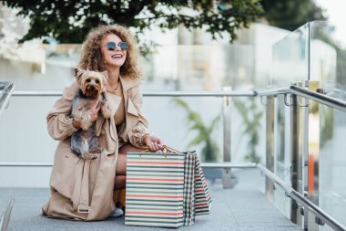 Vitrinlere bakan bir kadın, dükkanın yakınında oturan bir kadın. İçeri girmeden önce gülümseyen kadın dükkanın camını işaret ediyor. Genç bir kadın köpeğiyle birlikte vitrindeki kıyafetlere bakıyor..