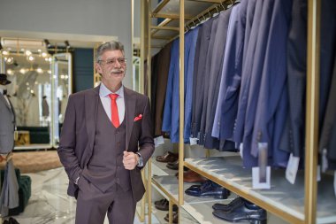 Muhteşem milyoner portresi ya da zengin yetişkin bir adam lüks moda butiğini ziyaret ederek özel bir kostüm alıyor. Arka planda hazır takım elbise askısı..