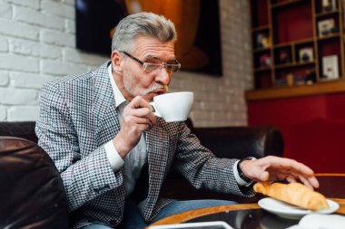 Orta yaşlı, şık takım elbiseli bir adam bir fincan kahve ve kruvasanla iyi bir hafta sonu geçirsin..