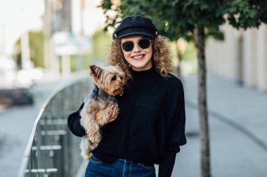 Uzun kıvırcık saçlı mutlu kadın küçük köpeği tutuyor. Güzel kız küçük köpeğe sarılıyor. Köpekli kadın. Yorkshire Teriyeri 'yle gülümseyen çekici kadın. Ellerinde köpek olan bir kız. Evcil hayvan evlat edinme, evcil hayvan hayatı.