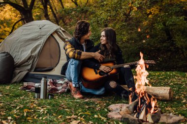 Genç bir çift turist, doğadaki şenlik ateşinin yanında dinleniyor. Yakışıklı adam kızına kamp ateşinin yanında yürüyüş yaparken gitar çalmayı öğretiyor. Turistik konsept resmi.
