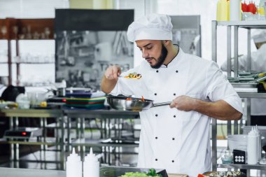 Beyaz üniformalı, gülümseyen genç şef çeşitli sebzelerle yemek hazırlıyor..