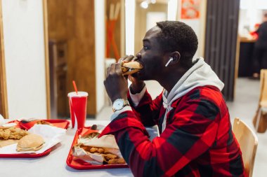 Afro-Amerikalı bir adam hamburgerciyi ısırıyor. Yakışıklı ve afro tişörtlü genç bir adam. Bir fast food kafede hamburger tutuyor..