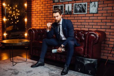 Şık bir adam elinde dergiyle gözlüğünü tutarken lüks bir dairede kanepede oturuyor..