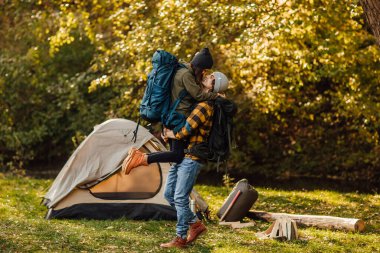 Ormanda çadırın yanında sırt çantalı genç ve güzel bir çift öpüşüyor. Çekici kadın ve yakışıklı erkek doğada birlikte dinleniyorlar. Instagram ruh hâli fotoğrafları. Yürüyüş sırasında insanlara sarılmak.
