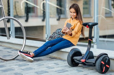 Mutlu kız çocuklar ellerinde telefonla güneşli bir yaz gününde parkta uçan kaykay sürmeyi öğrenirler..