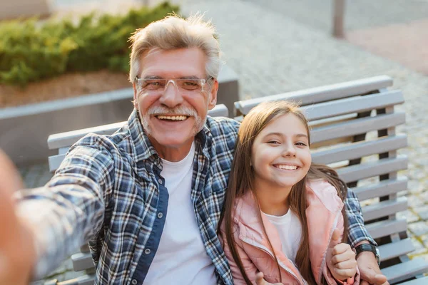 Foto Abuelo Una Nieta Sonrientes Sentados Banco Haciendo Una Selfie — Foto de Stock