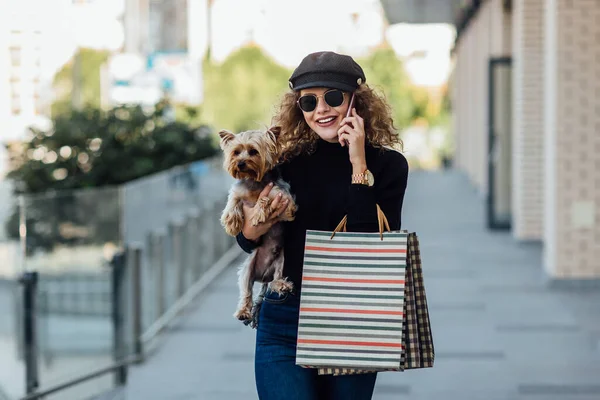 Fashion walk woman with long curly hair holds small dog and shopping bags. Beautiful girl hugs little dog. Smiling attractive woman with Yorkshire terrier. Girl with dog in hands, and sale concept. Business time.