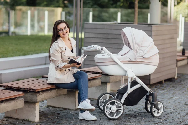 Lächelnde Hübsche Mutter Liest Buch Auf Bank Neben Kinderwagen Park — Stockfoto