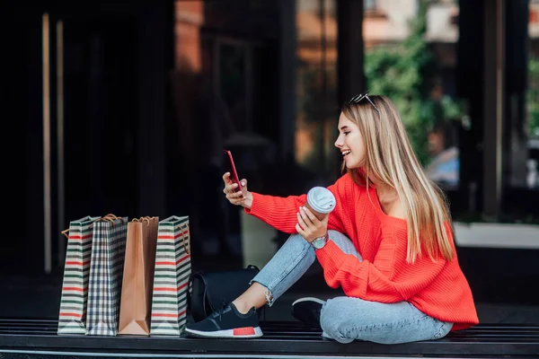 Piękna Blondynka Siedząca Ulicy Otoczona Torbami Zakupy Patrząca Telefon — Zdjęcie stockowe