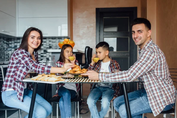 Family laughing around breakfast in kitchen while eating and children drinking fresh juice.