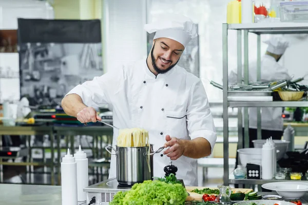 Bearded, professional chief cook in white unifirm, boil spahhetti, kitchen equipment.