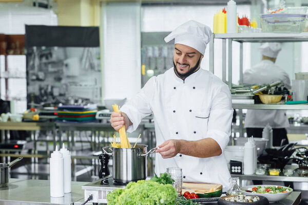 Portrait of chef with hat preparing delicious meal in professional kitchen surrounded by equipment. Stainless steel kitchen with spaghetti.
