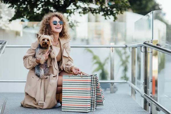 Vitrinlere bakan bir kadın, dükkanın yakınında oturan bir kadın. İçeri girmeden önce gülümseyen kadın dükkanın camını işaret ediyor. Genç bir kadın köpeğiyle birlikte vitrindeki kıyafetlere bakıyor..