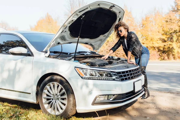 Young stressed woman driver standing near broked car with open hood.