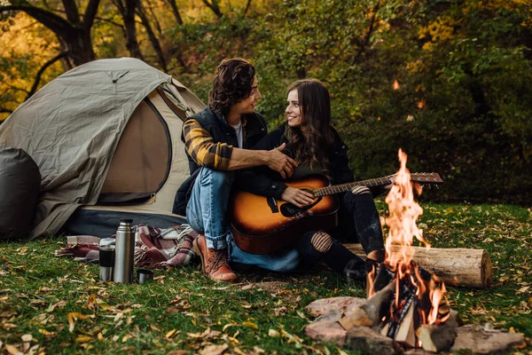 Young loving couple of tourists relaxing near the bonfire in the nature. Handsome man teaches his girl to play guitar on a hike near the campfire. Touristic concept picture.