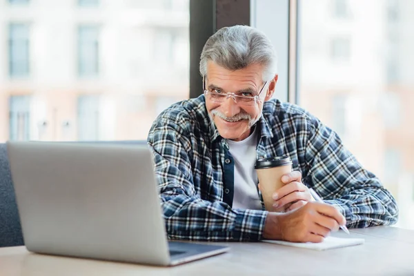 Positive old businessman working on laptop computer in cafe drinking coffee, lifestyle concept.
