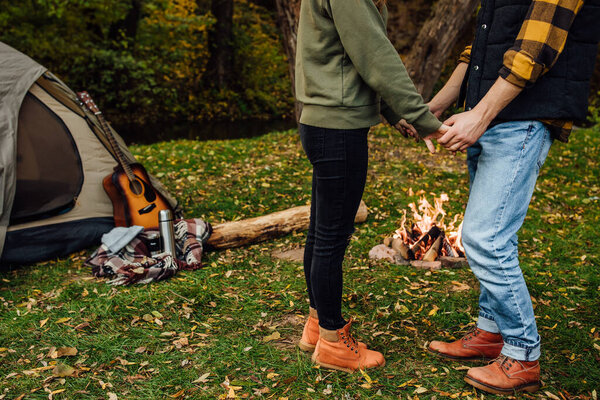 Close up photo of handsome man and attrective woman holding hands. Young loving couple of tourists relaxing near the bonfire in the nature.