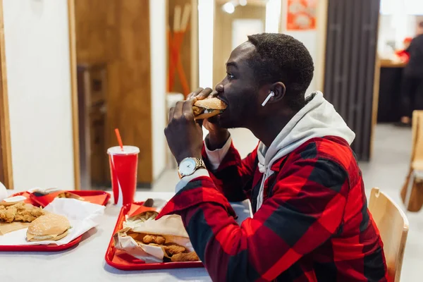 Afro-Amerikalı bir adam hamburgerciyi ısırıyor. Yakışıklı ve afro tişörtlü genç bir adam. Bir fast food kafede hamburger tutuyor..