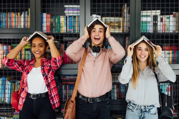 International friendship, fun and teenage concept. Group of three cheerful students teenagers in casual outfits with books in the form of a roof over the head fooling around in library.