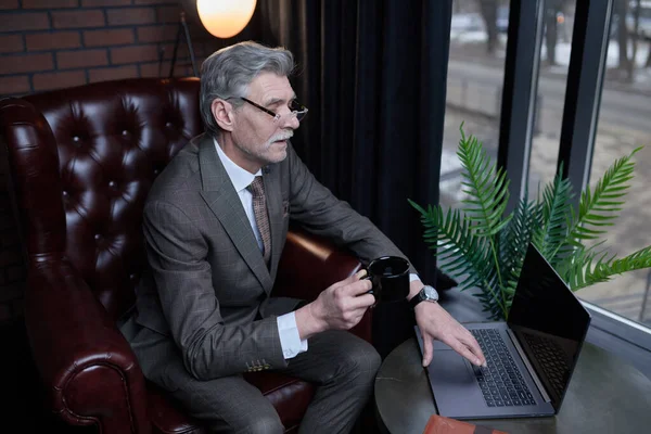 Old millionaire in business suit with cup of coffee in hands solving business issues on the laptop of stylish office.