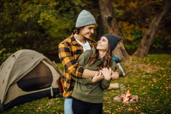 Portrait of happy loving couple of tourist in casual clothes in the forest near tent. A handsome man hugs an attractive woman looking face to face.