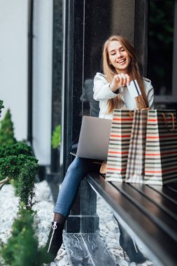 Yeni kıyafetler aldıktan sonra sarışın bir kız alışveriş merkezinin yanında oturur ve kredi kartıyla online alışveriş yapar..