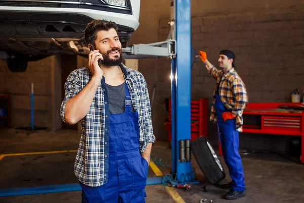 Uśmiechnięty Mechanik Rozmawiający Smartfonie Podnoszonym Samochodem Współpracownikiem Tle — Zdjęcie stockowe