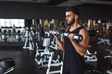 Spor salonundaki kaslı genç adam, pazılarında dambıllarla egzersiz yapıyor..