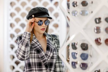 Göz kamaştırıcı sarışın kadın, optik mağazada gözlüklü rafların yanında güneş gözlüğü deniyor..