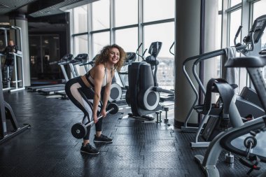 Sarışın bir fitness kızı canlandırıcıların yanındaki spor salonunda halterle çömelirken portresi..