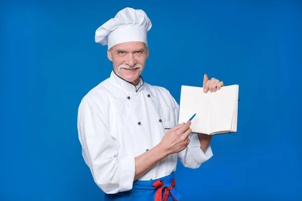 新しいレシピを探して 彼のメモ帳で 青の背景に隔離された高齢者のマスターシェフの写真 — ストック写真